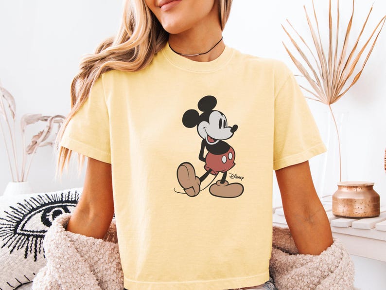 以下が含まれることがあります： ミッキーマウスのイラストが描かれた、明るい黄色のTシャツ。漫画のキャラクターは、赤いショートパンツ、白い手袋、茶色の靴というクラシックな衣装で描かれています。ディズニーのロゴが見えます。