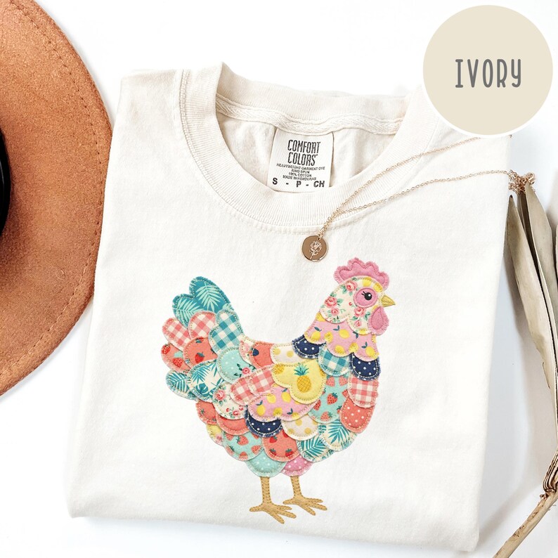 Op de afbeelding: Ivoor kleurig T-shirt met een kleurrijk patchwork kippenontwerp. De kip heeft verschillende patronen, waaronder bloemen-, ruit- en fruitprints. Het shirt heeft een ronde hals en korte mouwen. Een gouden ketting met een hanger is over het shirt gedrapeerd.