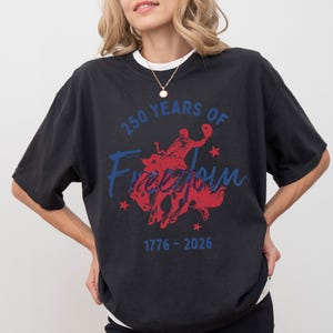 May include: Black t-shirt with a graphic design celebrating 250 years of freedom. The design features a red silhouette of a person riding a bucking bronco, with blue text that reads "250 Years of Freedom" and "1776-2026".