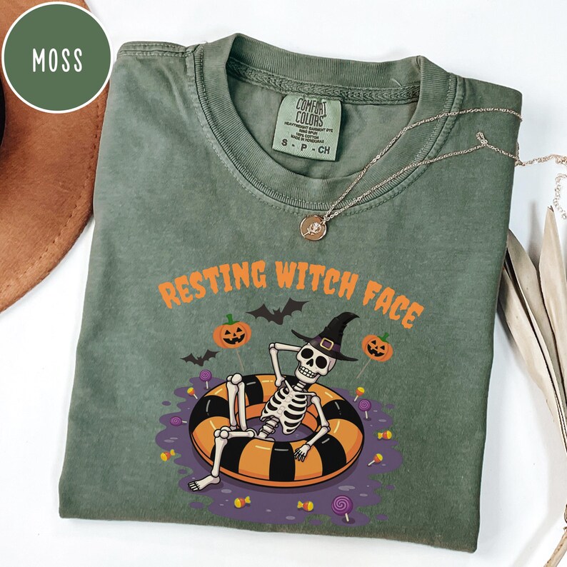 May include: A moss green t-shirt with the words "RESTING WITCH FACE" in orange. The shirt features a skeleton wearing a witch hat and floating in an orange and black inner tube. Pumpkins, bats, and candy complete the Halloween design.
