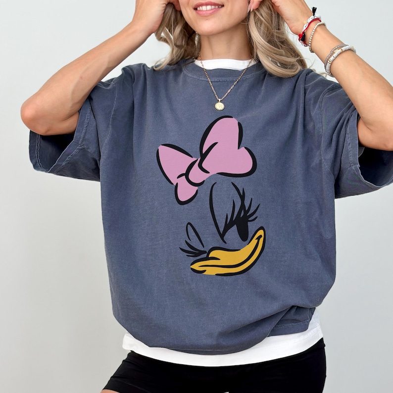 May include: A blue-grey t-shirt featuring a cartoon graphic of Daisy Duck's face. The graphic includes a pink bow, black eyelashes, and a yellow beak. The shirt has short sleeves and a white undershirt.