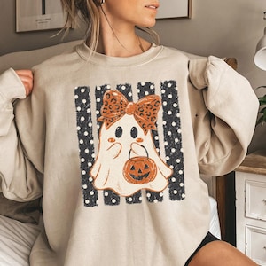 Puede incluir: Una sudadera beige de cuello redondo con el gráfico de un fantasma blanco que lleva un lazo con estampado de leopardo y sostiene un cubo de calabaza naranja. El fantasma está centrado sobre un fondo de rayas verticales negras con lunares blancos.