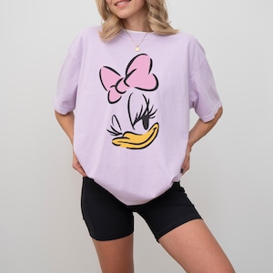 May include: A light purple t-shirt featuring a cartoon graphic of a character's face with a pink bow, black eyelashes, and a yellow beak. The shirt is paired with black shorts.