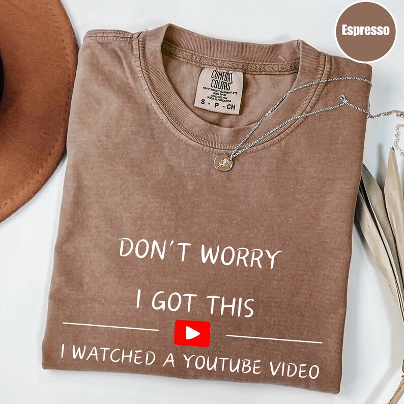 May include: Espresso-colored t-shirt with the text "DON'T WORRY I GOT THIS I WATCHED A YOUTUBE VIDEO" in white. The shirt has a small YouTube play button graphic. A silver necklace with a pendant is draped across the shirt.