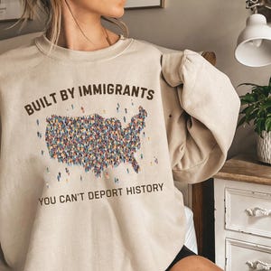 May include: Beige sweatshirt with the text "BUILT BY IMMIGRANTS" above a graphic of the United States filled with tiny figures. Below the graphic, the text reads "YOU CAN'T DEPORT HISTORY".
