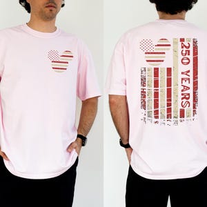 May include: A pale pink t-shirt with a Mickey Mouse-themed design in red and white stripes and dots. The back of the shirt has a larger design with the text "250 YEARS" in red. The shirt is worn by a person.