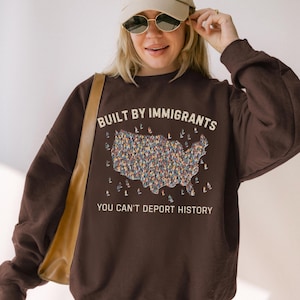 May include: A brown sweatshirt with the text "BUILT BY IMMIGRANTS" above a colorful illustration of the United States filled with people. Below the illustration is the text "YOU CAN'T DEPORT HISTORY". The person in the image is wearing a beige cap and sunglasses.