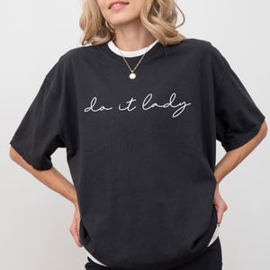 Puede incluir: Camiseta negra con el texto manuscrito "do it lady" en escritura blanca. La camiseta tiene cuello redondo y mangas cortas. La modelo lleva un collar dorado con un colgante.