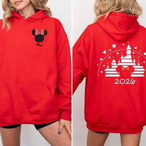 Puede incluir: Una sudadera con capucha roja con un bolsillo delantero y un dise&ntilde;o de Minnie Mouse. La parte delantera presenta una silueta negra de Minnie Mouse con un lazo de lunares rojos y blancos y la palabra "Mam&aacute;". La parte trasera muestra un dise&ntilde;o de castillo blanco con el a&ntilde;o "2026".