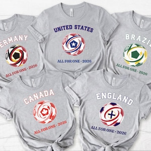 May include: Five gray t-shirts with soccer ball graphics representing Germany, United States, Brazil, Canada, and England. Each shirt has the country's name and "ALL FOR ONE - 2026" printed on it. The soccer balls feature the colors of each country's flag.