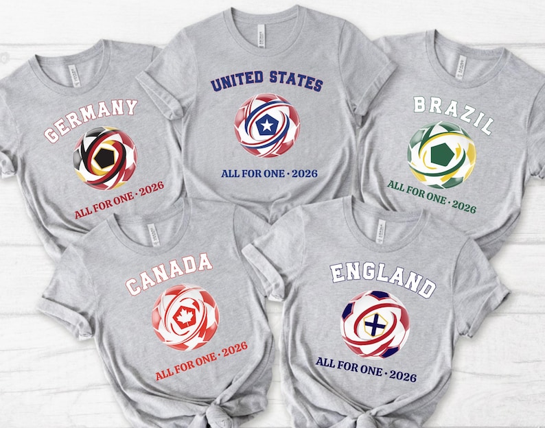 May include: Five gray t-shirts with soccer ball graphics representing Germany, United States, Brazil, Canada, and England. Each shirt has the country's name and "ALL FOR ONE - 2026" printed on it. The soccer balls feature the colors of each country's flag.