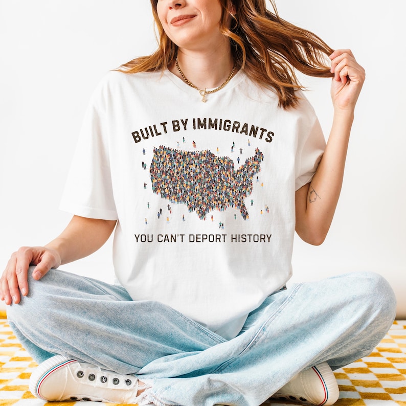 May include: White t-shirt with the text "BUILT BY IMMIGRANTS" above a map of the United States filled with tiny figures. Below the map, the text reads "YOU CAN'T DEPORT HISTORY". The person is wearing light blue jeans.