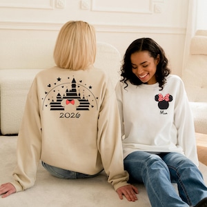 Puede incluir: Dos sudaderas de manga larga. Una es beige con un dise&ntilde;o de castillo negro y el a&ntilde;o 2026. La otra es blanca con un dise&ntilde;o de Minnie Mouse y la palabra "Mam&aacute;". Ambas son usadas por personas sentadas en una superficie blanca.