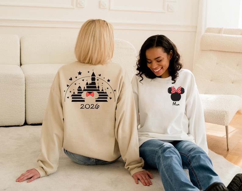 Puede incluir: Dos sudaderas de manga larga. Una es beige con un dise&ntilde;o de castillo negro y el a&ntilde;o 2026. La otra es blanca con un dise&ntilde;o de Minnie Mouse y la palabra "Mam&aacute;". Ambas son usadas por personas sentadas en una superficie blanca.