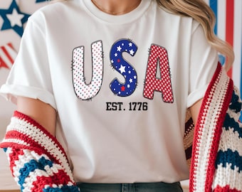 May include: White t-shirt with the word "USA" in red, white, and blue, with the text "EST. 1776" below. The "U" is white with red and blue polka dots, the "S" is blue with white stars, and the "A" is red with white stars. The shirt is part of a patriotic theme.