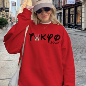 May include: A red sweatshirt with the word "TOKYO" in black script, featuring a Minnie Mouse graphic and the year "2026". The image also shows a beige cap with text and sunglasses.