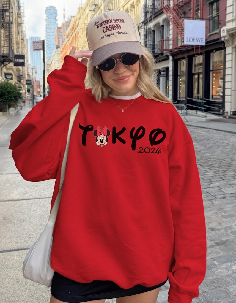 May include: A red sweatshirt with the word "TOKYO" in black script, featuring a Minnie Mouse graphic and the year "2026". The image also shows a beige cap with text and sunglasses.