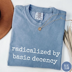 Könnte beinhalten: Ein blaues Jeans-T-Shirt mit dem weißen Aufdruck "radicalized by basic decency". Eine goldene Halskette mit Anhänger liegt über dem Shirt. Das Shirt ist gefaltet und hat ein Etikett mit der Aufschrift "Comfort Colors".