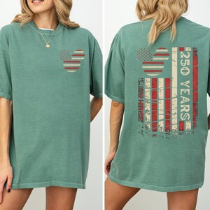 May include: A sage green t-shirt featuring a Mickey Mouse head design on the front and back. The design incorporates red and white stripes and stars, with the text "250 YEARS" on the back, in a vintage style.