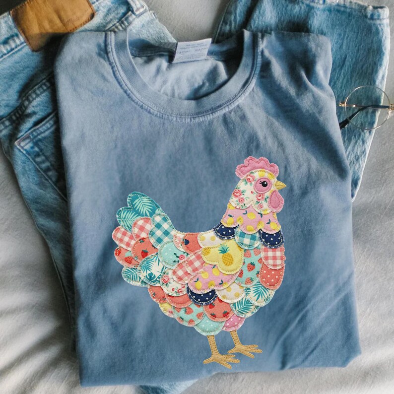 Op de afbeelding: Een lichtblauw T-shirt met een kleurrijk patchwork kippenontwerp. De kip heeft een roze kam en lellen, met een lichaam gemaakt van verschillende stoffen met patronen, waaronder ruitjes, bloemen en fruitprints. Het shirt wordt gecombineerd met een blauwe jeans.