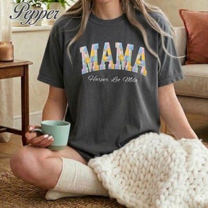 Puede incluir: Camiseta gris oscuro con la palabra "MAMA" en letras de patchwork coloridas. Debajo de la palabra "MAMA" está el texto "Harper Leo Mila". La persona sostiene una taza verde claro y lleva calcetines blancos.