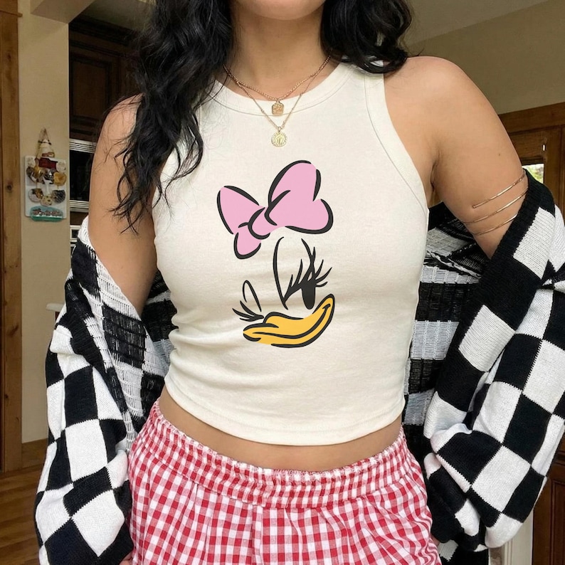 Op de afbeelding: Cr&egrave;mekleurige tanktop met een cartoonafbeelding van een eend met een roze strik en gele snavel. De top wordt gecombineerd met een rood-wit geruite pyjamabroek en een zwart-wit geruit vest.