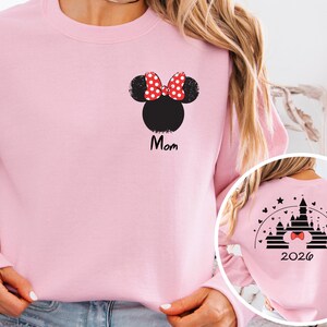 Puede incluir: Sudadera rosa claro con una cabeza de Minnie Mouse negra y un lazo de lunares rojos y blancos. La palabra "Mom" est&aacute; impresa en negro debajo. La parte trasera de la sudadera tiene un dise&ntilde;o de castillo y el a&ntilde;o 2026.