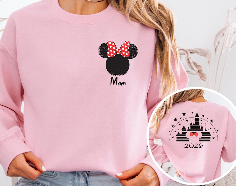 Puede incluir: Sudadera rosa claro con una cabeza de Minnie Mouse negra y un lazo de lunares rojos y blancos. La palabra "Mom" est&aacute; impresa en negro debajo. La parte trasera de la sudadera tiene un dise&ntilde;o de castillo y el a&ntilde;o 2026.