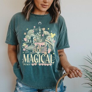 May include: A dark teal short-sleeve t-shirt featuring a graphic print with a castle, monorail, and various park icons. The text reads The Most MAGICAL PLACE in cream and pink lettering, surrounded by small flowers and heart shapes.