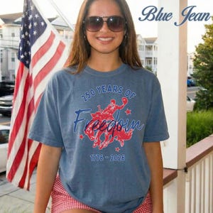 May include: Blue short-sleeved t-shirt with the text "250 Years of Freedom" and a red graphic of a person riding a horse. The shirt also has the years 1776-2026 printed on it. An American flag is in the background.