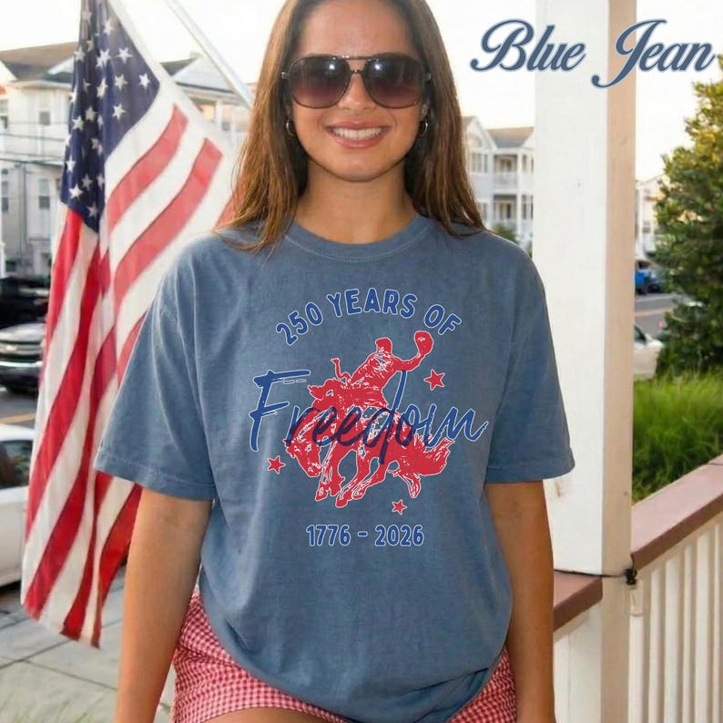 May include: Blue short-sleeved t-shirt with the text "250 Years of Freedom" and a red graphic of a person riding a horse. The shirt also has the years 1776-2026 printed on it. An American flag is in the background.