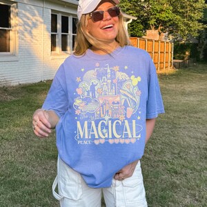 May include: A periwinkle blue short-sleeve t-shirt featuring a graphic print with the text The Most Magical Place. The design includes illustrations of a castle, a monorail, teacups, and various floral and star accents in yellow, pink, and white.