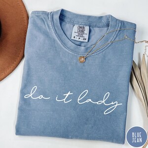 Puede incluir: Una camiseta azul vaquero Comfort Colors con la frase "do it lady" en escritura blanca. Un collar dorado con un colgante descansa sobre la camisa. La camisa est&aacute; doblada y colocada sobre una superficie blanca.