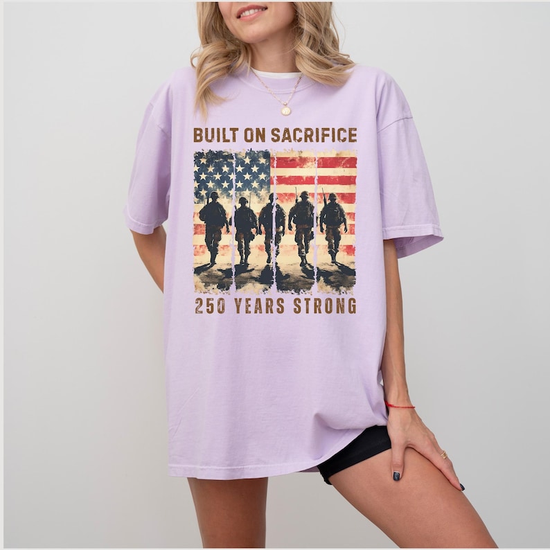 May include: A light purple t-shirt featuring an American flag design with silhouettes of soldiers and the text "BUILT ON SACRIFICE" and "250 YEARS STRONG". The shirt has short sleeves and a relaxed fit.