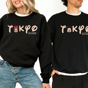 May include: Black sweatshirts with the word "TOKYO" in a stylized font, featuring Mickey and Minnie Mouse characters. The year "2026" is below the word. The sweatshirts have a crew neck and long sleeves.