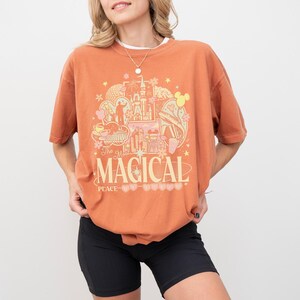 May include: A burnt orange short-sleeve t-shirt featuring a graphic print with various theme park icons, stars, and flowers. The text reads "The Most MAGICAL PLACE ON EARTH" in a retro-style font. The shirt is paired with black athletic shorts.