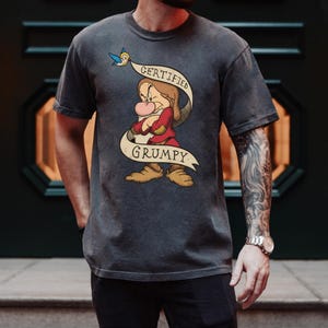 May include: Dark gray t-shirt featuring a cartoon image of Grumpy from Snow White. The character is depicted with crossed arms, a red shirt, and the words "CERTIFIED GRUMPY" on a banner. A blue bird is perched above the banner.