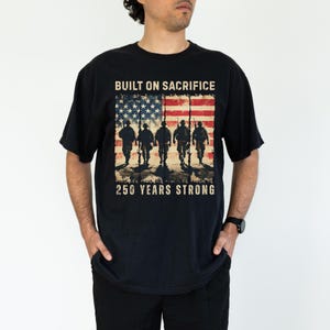 May include: Black t-shirt featuring an American flag design with soldiers and the text "BUILT ON SACRIFICE" and "250 YEARS STRONG." The graphic is distressed, giving a vintage look.
