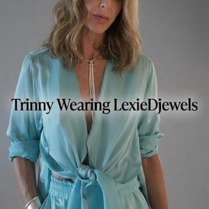 May include: A light blue satin button-down shirt tied at the waist with matching elastic-waist trousers, a thin white cord necklace, and wide silver cuff bracelets on both wrists. The text "Trinny Wearing LexieDjewels" is centred on the image.