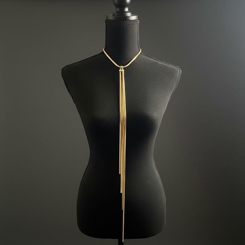 May include: A gold-colored necklace with a Y-shaped design. The necklace features a delicate chain that splits into multiple strands, creating a cascading effect. The necklace is displayed on a black mannequin, against a dark background.
