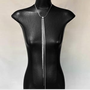 May include: A silver-toned necklace with a long, vertical design. The necklace features multiple strands that cascade down from a central point, creating a sleek and modern look. The jewelry is displayed on a black mannequin.