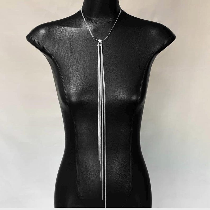 May include: A silver-toned necklace with a long, vertical design. The necklace features multiple strands that cascade down from a central point, creating a sleek and modern look. The jewelry is displayed on a black mannequin.