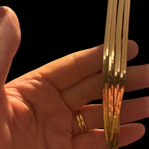 May include: A hand holding multiple strands of gold-colored, flat herringbone chains. The chains are arranged in a cascading pattern, reflecting light. A gold ring is on the finger. The background is black.