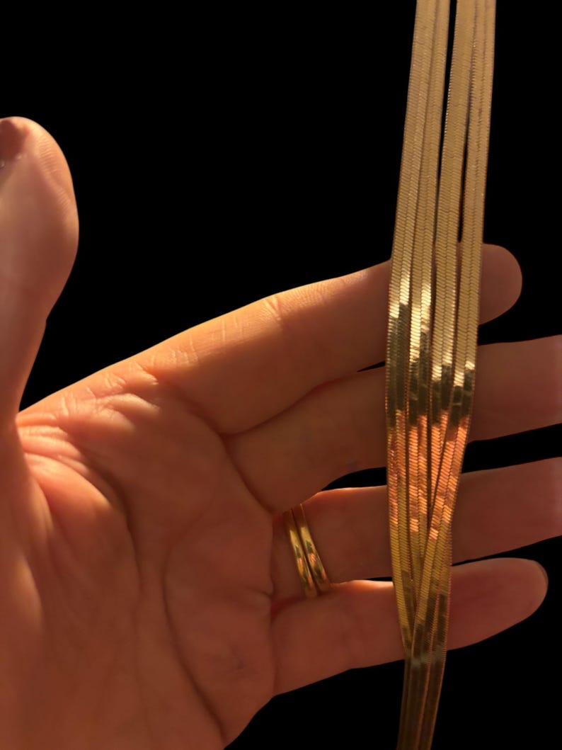 May include: A hand holding multiple strands of gold-colored, flat herringbone chains. The chains are arranged in a cascading pattern, reflecting light. A gold ring is on the finger. The background is black.