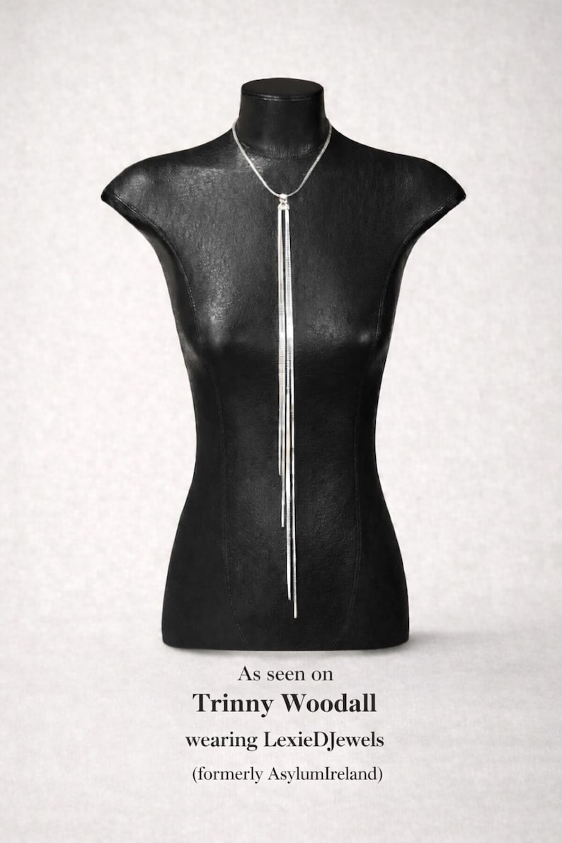 May include: A silver necklace with a vertical bar pendant, displayed on a black mannequin. The necklace has a delicate chain and three long, slender bars that hang down. The mannequin is on a white background.