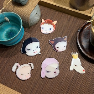 May include: A collection of six acrylic pins featuring stylized cartoon character faces. The designs include a cat-eared figure, a character with black hair, a dark-eared creature, a rabbit-eared figure, a person in a pink hood, and a white dog with a crown.