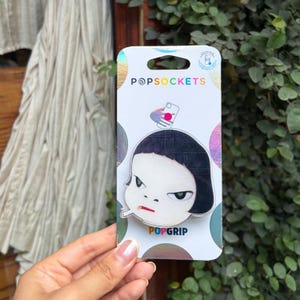 May include: A PopSockets PopGrip phone accessory featuring a graphic of a stylized character with black hair, green eyes, and a cigarette in their mouth. The product is mounted on a white cardboard backing with the text PopSockets and PopGrip.
