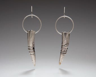 May include: A pair of silver-toned drop earrings featuring a circular hoop at the top and a textured, tapered, horn-shaped pendant suspended from the hoop. The pendant is wrapped with thin silver wire around the lower section.