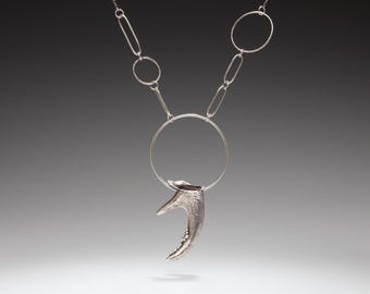 May include: A silver necklace featuring a large circular pendant attached to a textured, curved, horn-like element. The chain consists of alternating thin circular and elongated oval links, creating a geometric design with a metallic finish.