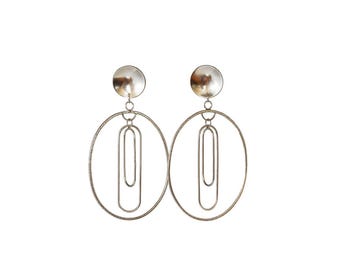 May include: A pair of silver-toned drop earrings featuring a circular stud top connected to a large oval hoop. Inside each large hoop, two smaller, elongated oval wire links hang vertically, creating a layered, geometric, and dangling design.